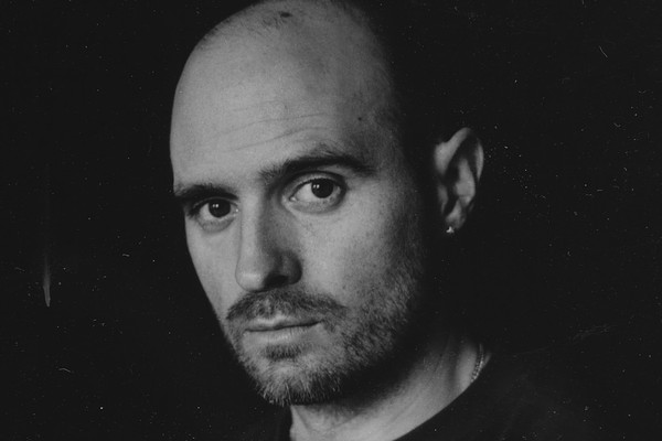 A close-up black and white photo shows artist Nick Schofield in a black t-shirt against a black background looking directly at the camera. 