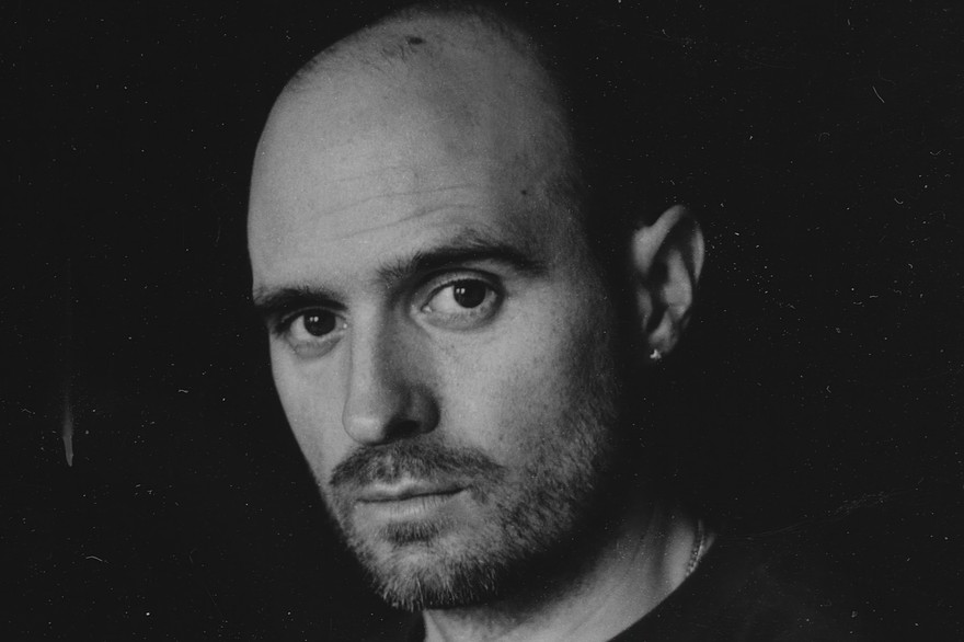A close-up black and white photo shows artist Nick Schofield in a black t-shirt against a black background looking directly at the camera. 