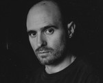 A close-up black and white photo shows artist Nick Schofield in a black t-shirt against a black background looking directly at the camera. 