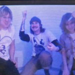 Three members of NIFE FITE pose together against a light‑colored wall, captured in a bluish, retro‑styled photo. One crouches holding an object, another kneels with a beanie and raised arm, and the third stands leaning in while holding something near their head, creating an energetic, chaotic group portrait.