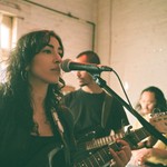 Artist Niloo is seen playing guitar and singing in a bright room with white brick walls and a draped window in the background. Two other members are seen behind them. 