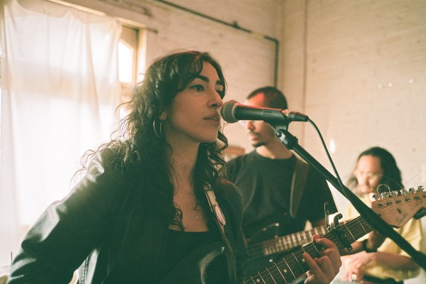 Artist Niloo is seen playing guitar and singing in a bright room with white brick walls and a draped window in the background. Two other members are seen behind them. 