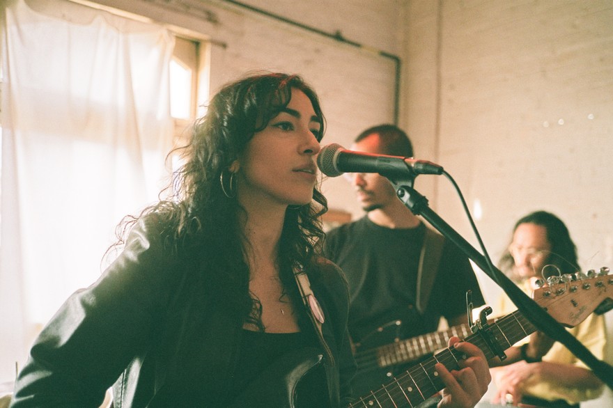 Artist Niloo is seen playing guitar and singing in a bright room with white brick walls and a draped window in the background. Two other members are seen behind them. 
