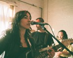Artist Niloo is seen playing guitar and singing in a bright room with white brick walls and a draped window in the background. Two other members are seen behind them. 