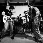 A black sand white photo shows three-piece rock band Often Wrong performing onstage. The guitar player and bassist are seen in the foreground, with the drummer in the middle in the background. 