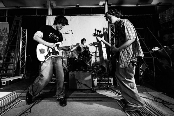 A black sand white photo shows three-piece rock band Often Wrong performing onstage. The guitar player and bassist are seen in the foreground, with the drummer in the middle in the background. 