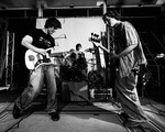 A black sand white photo shows three-piece rock band Often Wrong performing onstage. The guitar player and bassist are seen in the foreground, with the drummer in the middle in the background. 