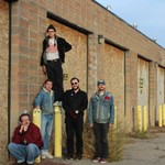 Five members of the band NON ULTRAS are seen standing in an old alleway against a brick building. One member in the middle is seen standing on top of a yellow concrete post. 