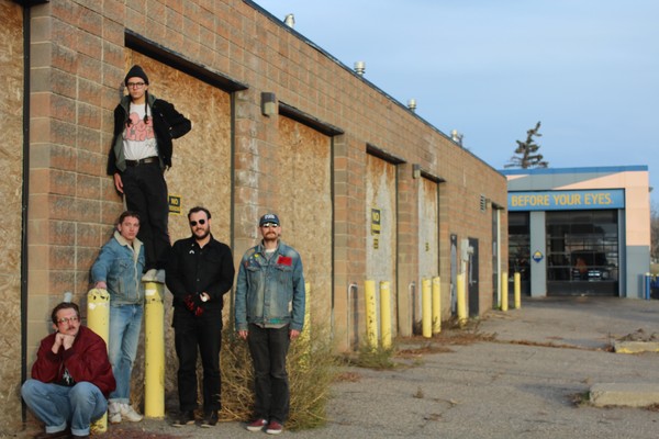 Five members of the band NON ULTRAS are seen standing in an old alleway against a brick building. One member in the middle is seen standing on top of a yellow concrete post. 