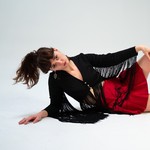 A colour photo shows Nora Kelly against an off-white background laying on their side and looking at the camera. They are wearing a bright red skirt, and a black tassled button top.