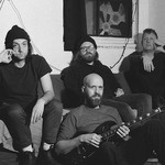 A grainy black and white photo shows four members of the group Oil Giant sitting in a living room. Three members sit on the couch, and one member sits on the floor in front holding an electric guitar. 