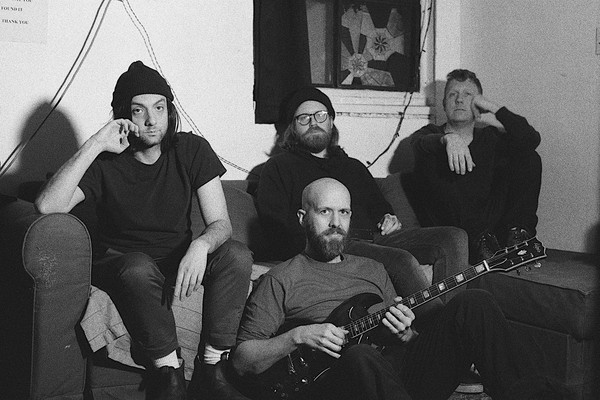 A grainy black and white photo shows four members of the group Oil Giant sitting in a living room. Three members sit on the couch, and one member sits on the floor in front holding an electric guitar. 