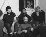 A grainy black and white photo shows four members of the group Oil Giant sitting in a living room. Three members sit on the couch, and one member sits on the floor in front holding an electric guitar. 