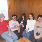 Five members of Our Friend, Oscar sit close together on a couch in a wood‑paneled room, casually dressed and relaxed. A whiteboard behind them lists handwritten song titles, hinting at a rehearsal or planning session, while the group smiles toward the camera in a warm, collaborative setting.