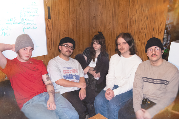 Five members of Our Friend, Oscar sit close together on a couch in a wood‑paneled room, casually dressed and relaxed. A whiteboard behind them lists handwritten song titles, hinting at a rehearsal or planning session, while the group smiles toward the camera in a warm, collaborative setting.