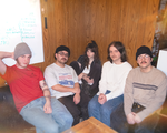 Five members of Our Friend, Oscar sit close together on a couch in a wood‑paneled room, casually dressed and relaxed. A whiteboard behind them lists handwritten song titles, hinting at a rehearsal or planning session, while the group smiles toward the camera in a warm, collaborative setting.