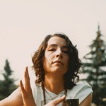 A colour photo shows the frontperson of the project Old Mound sitting outside in a grassy field with pine trees in the background. They have brown hair, a white outfit, and are seen holding a small metal chalice. 