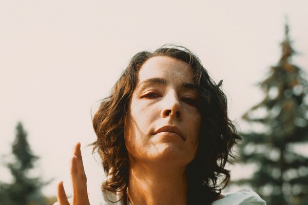 A colour photo shows the frontperson of the project Old Mound sitting outside in a grassy field with pine trees in the background. They have brown hair, a white outfit, and are seen holding a small metal chalice. 