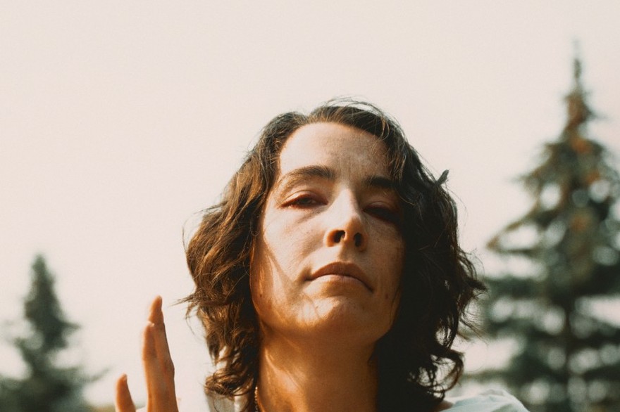 A colour photo shows the frontperson of the project Old Mound sitting outside in a grassy field with pine trees in the background. They have brown hair, a white outfit, and are seen holding a small metal chalice. 