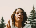 A colour photo shows the frontperson of the project Old Mound sitting outside in a grassy field with pine trees in the background. They have brown hair, a white outfit, and are seen holding a small metal chalice. 