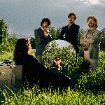 A colour photo shows the band Oranje outside in a green field with long grass and bushes. Three members are seen wearing blazers and facing the camera. Another member sits with their back to the camera, looking into a mirror with a corded phone to their ear, and a filing cabinet sitting to their left. 