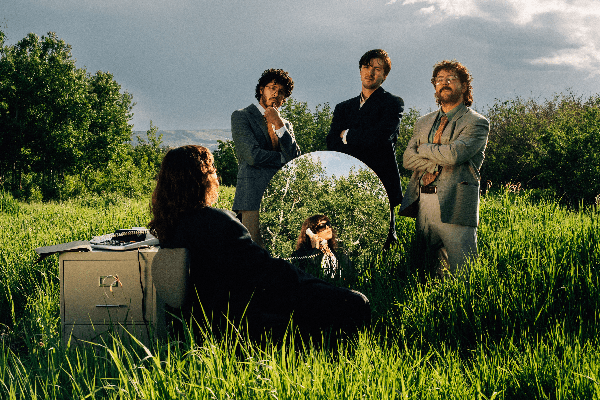 A colour photo shows the band Oranje outside in a green field with long grass and bushes. Three members are seen wearing blazers and facing the camera. Another member sits with their back to the camera, looking into a mirror with a corded phone to their ear, and a filing cabinet sitting to their left. 