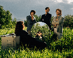 A colour photo shows the band Oranje outside in a green field with long grass and bushes. Three members are seen wearing blazers and facing the camera. Another member sits with their back to the camera, looking into a mirror with a corded phone to their ear, and a filing cabinet sitting to their left. 