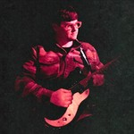 A colour photo shows the frontperson of band Pearly Moon bathed in red light standing and playing guitar, fading into a black background. 