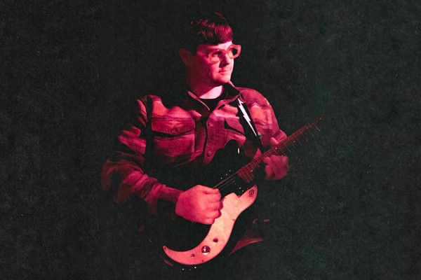 A colour photo shows the frontperson of band Pearly Moon bathed in red light standing and playing guitar, fading into a black background. 
