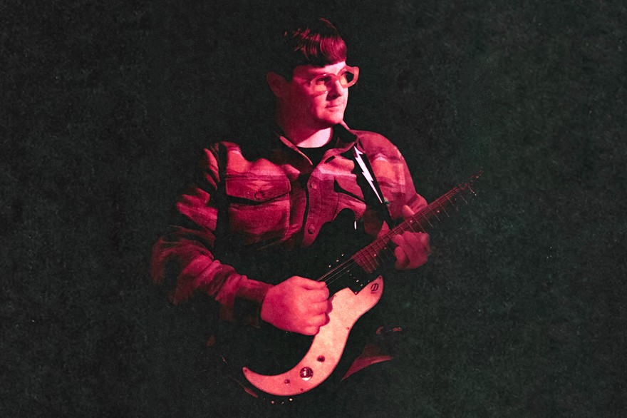 A colour photo shows the frontperson of band Pearly Moon bathed in red light standing and playing guitar, fading into a black background. 