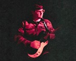 A colour photo shows the frontperson of band Pearly Moon bathed in red light standing and playing guitar, fading into a black background. 