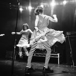 A black and white photo shows four members of the band PERRA performing on stage. The two in the middle are seen mid-dance, with the guitar player on their left, and the bassist on their right. 