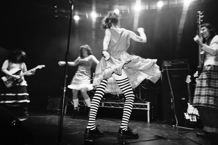 A black and white photo shows four members of the band PERRA performing on stage. The two in the middle are seen mid-dance, with the guitar player on their left, and the bassist on their right. 