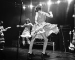 A black and white photo shows four members of the band PERRA performing on stage. The two in the middle are seen mid-dance, with the guitar player on their left, and the bassist on their right. 