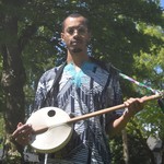 A colour photo shows artist Pesewa posing with a kologo instrument they have strapped on. They are outside with house and green trees seen behind them. 