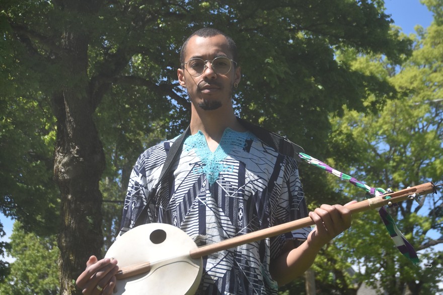 A colour photo shows artist Pesewa posing with a kologo instrument they have strapped on. They are outside with house and green trees seen behind them. 