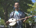 A colour photo shows artist Pesewa posing with a kologo instrument they have strapped on. They are outside with house and green trees seen behind them. 