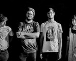 A black and white photo shows four-piece group Pike standing in a line with a dark black background behind them. 