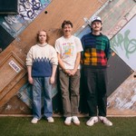A colour photo shows three-piece group Pinball, 1973 standed against a bizarre wall built out of various doors stacked diagonally. The group stands on a grassy patch in front of it. 