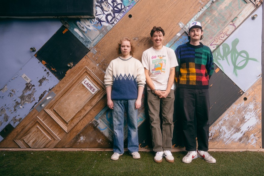 A colour photo shows three-piece group Pinball, 1973 standed against a bizarre wall built out of various doors stacked diagonally. The group stands on a grassy patch in front of it. 