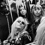 A black and white photo of the band Plezher shows them looking into a bathroom mirror covered with grafitti. The member in the far back is seen wearing a studded leather gimp mask. 