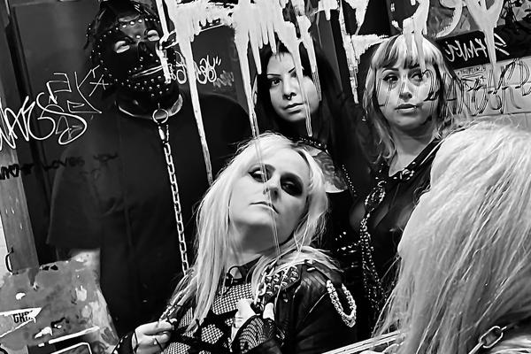 A black and white photo of the band Plezher shows them looking into a bathroom mirror covered with grafitti. The member in the far back is seen wearing a studded leather gimp mask. 