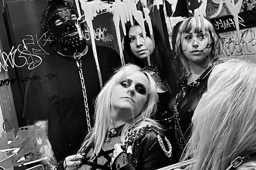 A black and white photo of the band Plezher shows them looking into a bathroom mirror covered with grafitti. The member in the far back is seen wearing a studded leather gimp mask. 