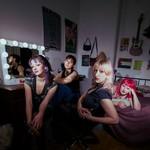 Four members of Pool Girl lounge together in a bedroom‑studio space filled with posters, a lit vanity mirror, and scattered personal objects. One reclines on the bed while the others sit on chairs or the floor, illuminated by dramatic lighting that highlights the room’s cozy, creative clutter.
