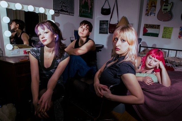 Four members of Pool Girl lounge together in a bedroom‑studio space filled with posters, a lit vanity mirror, and scattered personal objects. One reclines on the bed while the others sit on chairs or the floor, illuminated by dramatic lighting that highlights the room’s cozy, creative clutter.