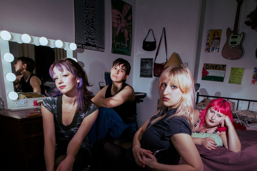 Four members of Pool Girl lounge together in a bedroom‑studio space filled with posters, a lit vanity mirror, and scattered personal objects. One reclines on the bed while the others sit on chairs or the floor, illuminated by dramatic lighting that highlights the room’s cozy, creative clutter.
