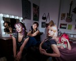 Four members of Pool Girl lounge together in a bedroom‑studio space filled with posters, a lit vanity mirror, and scattered personal objects. One reclines on the bed while the others sit on chairs or the floor, illuminated by dramatic lighting that highlights the room’s cozy, creative clutter.