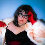 A colour photo shows the frontperson of project pseudo-antigone in front of a blue background wearing a white fur coat, red gloves, a black top and red lipstick. 