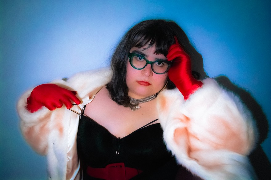 A colour photo shows the frontperson of project pseudo-antigone in front of a blue background wearing a white fur coat, red gloves, a black top and red lipstick. 