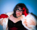 A colour photo shows the frontperson of project pseudo-antigone in front of a blue background wearing a white fur coat, red gloves, a black top and red lipstick. 
